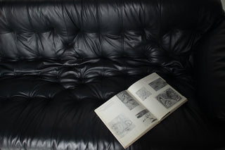 An open book with black-and-white sketches rests on a black tufted leather sofa.