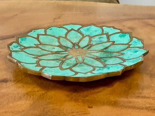 A round decorative tray with a turquoise and gold lotus flower pattern, resting on a wooden coffee table with a blurred sofa in the background.