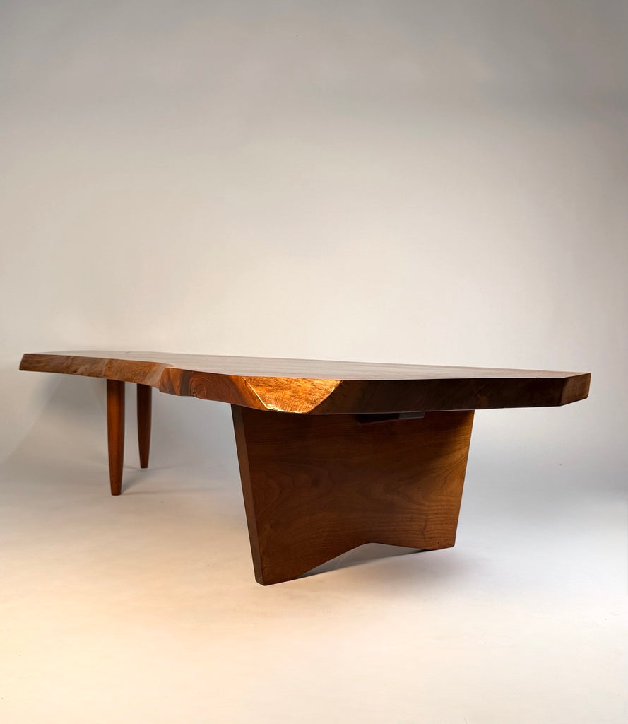 Profile of George Nakashima conoid coffee table showing the gently sloped English walnut tabletop the natural live edge as supported by sculptural leg base beneath.